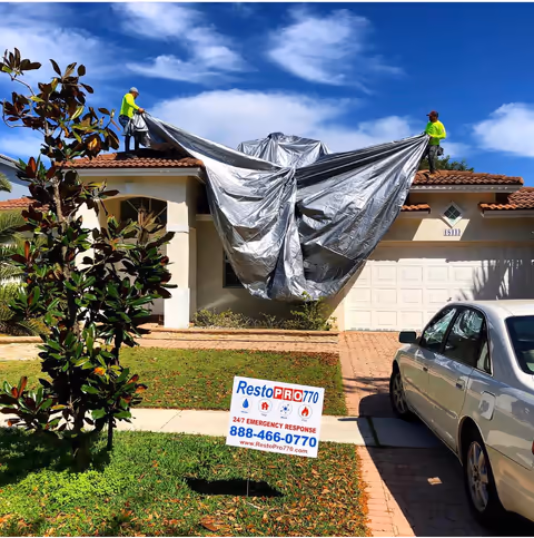 Two crew members unrolling a tarp over a house with a RestoPro770 sign in the yard